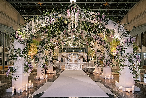 Ceremony aisle decor with a floral arch ceremony, white aisle runner, candles in glass cylinders, and hanging flowers in a high-ceiling hall