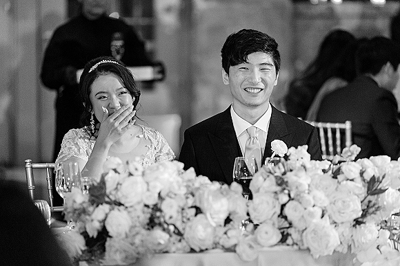 Reception couple moment at the wedding head table couple, bride laughing and covering her mouth as groom smiles in a black-and-white indoor venue