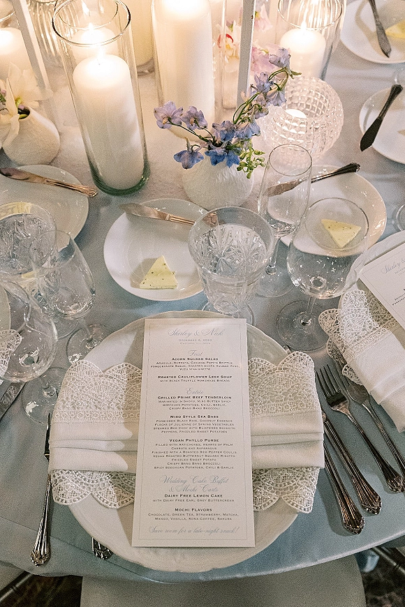 Reception tablescape with wedding place setting featuring menu card on white plates, lace napkins, blue flowers, and tall glass cylinder candles on light blue tablecloth