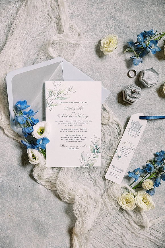 Invitation suite wedding invitation flat lay with envelope, vellum, blue ribbon, rings and earrings beside white roses on gray stone surface