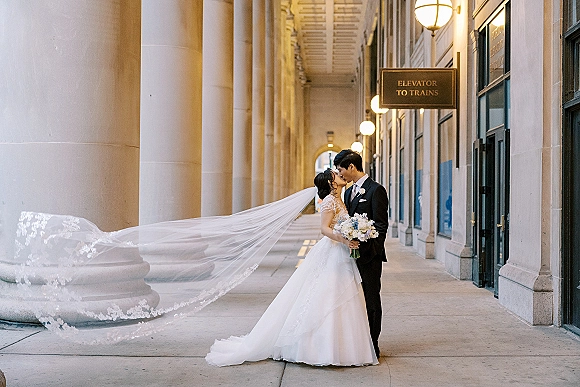 Wedding kiss portrait of bride and groom kissing, her cathedral veil flowing behind as she holds a bouquet on a columned city walkway