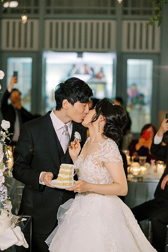 Cake cutting moment as bride and groom kiss, holding a cake plate and slice beside a tiered white wedding cake at a candlelit reception hall