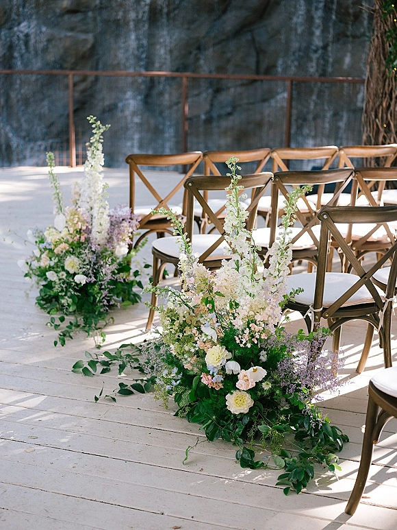 Ceremony aisle flowers in pastel clusters with greenery line a wood deck aisle beside cross back chairs and a stone wall backdrop