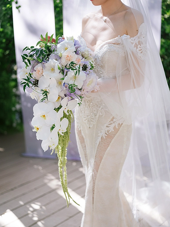 Bridal bouquet with cascading orchid bouquet of pastel blooms and greenery, held against a lace bodice and veil on a wooden deck outdoors