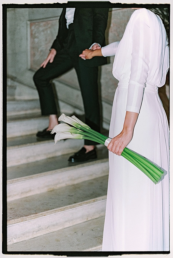 Wedding couple portrait of bride holding bouquet of white calla lilies as she and groom hold hands on stone stairs by a stone wall