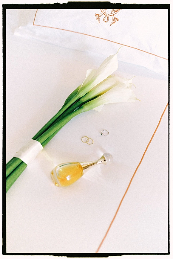 Wedding flat lay with calla lily bouquet, wedding rings, and perfume bottle on embroidered monogram linen over white fabric with gold trim border