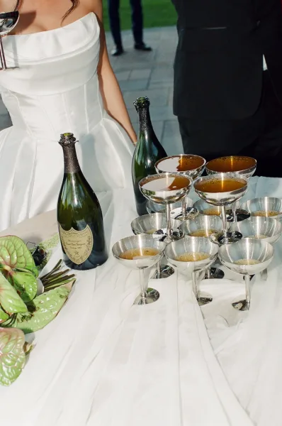 Champagne bar setup styled as a reception drink display, with coupe glasses arrayed on a bar table set on a stone patio.