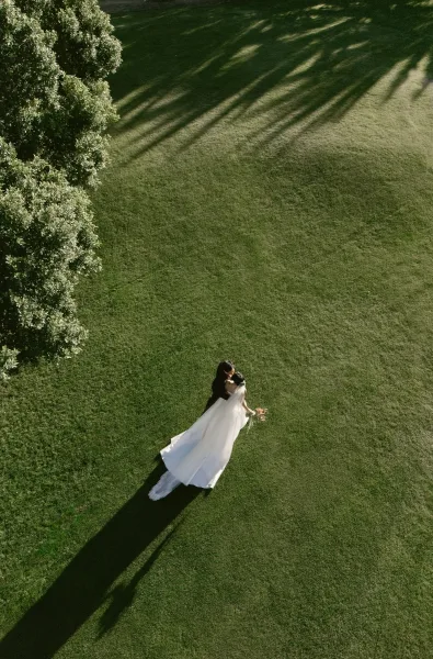 Couple portrait of the bride and groom embracing on a grass lawn beneath a leafy tree, her veil softly framing them as long shadows fall.