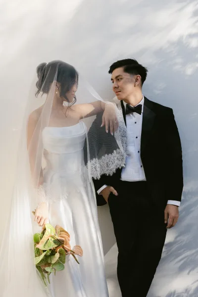 Couple portrait with the bride under veil, standing together against a white wall where leaf shadows create delicate patterns.