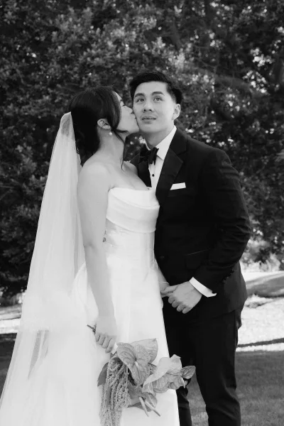 Wedding kiss portrait of a bride kissing her groom as she lifts a trailing bridal bouquet, framed by garden trees and lawn in soft afternoon light.