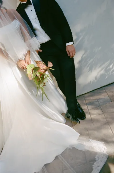 Couple portrait of a bride holding bouquet in an off-the-shoulder gown beside her groom, framed by dappled light on a white wall.