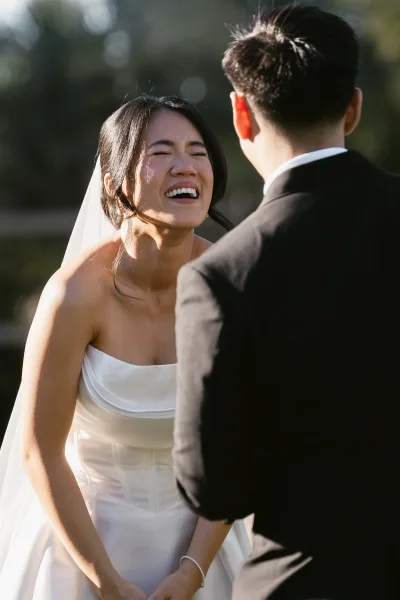Ceremony moment with a joyful wedding vows reaction as the bride laughs facing her groom in a black tuxedo beneath sunlit garden trees.
