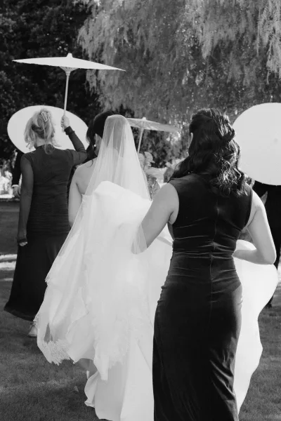 Ceremony processional with a bride walking with bridesmaids along a garden lawn, one shading her with a parasol as they near the ceremony.