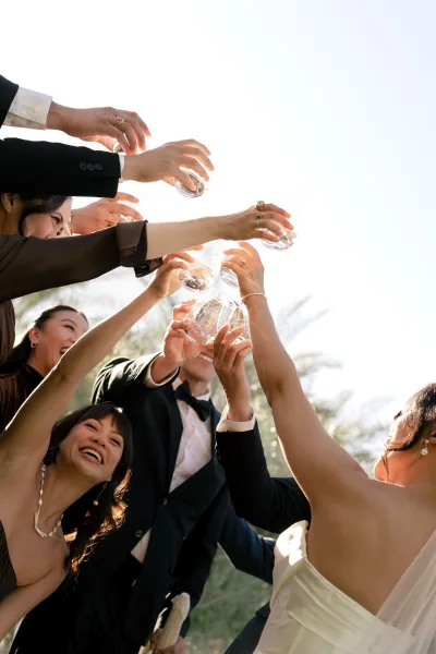 Wedding toast in a joyous group cheers moment, mixed glassware catching light as guests raise drinks under a blue sky framed by palm trees.