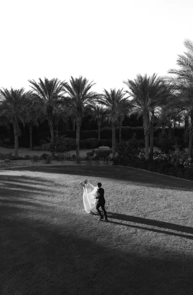 Bride and groom dancing in a couple portrait on a sunlit lawn, her veil flowing beneath tall palm trees and a clear sky