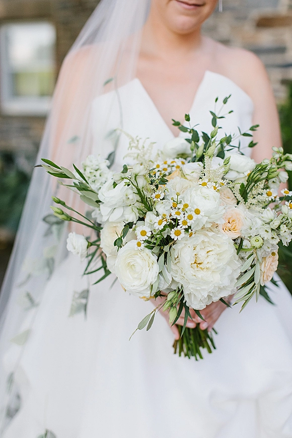 Bridal bouquet of white and green bridal bouquet with peonies, ranunculus, daisies and peach roses held against a stone wall window backdrop