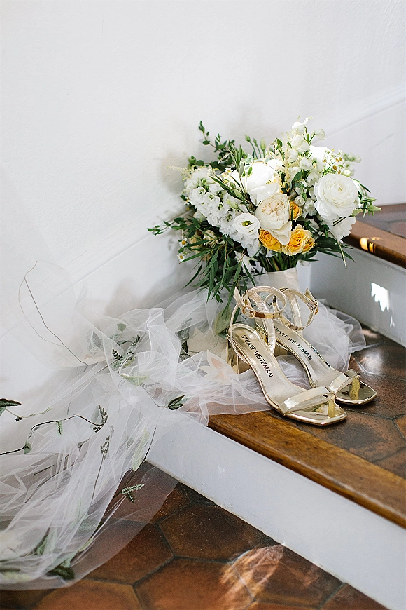 Bridal accessories flat lay with gold strappy heels beside a wedding bouquet and embroidered veil on a stair step against a white wall