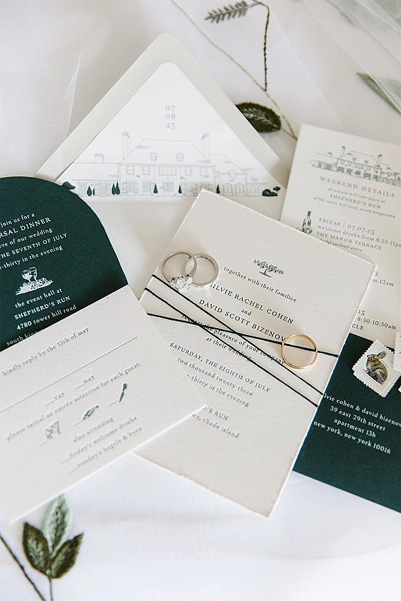 Wedding invitation suite invitation flat lay with rings, twine-tied envelope, venue illustration, RSVP and details cards on sheer floral fabric