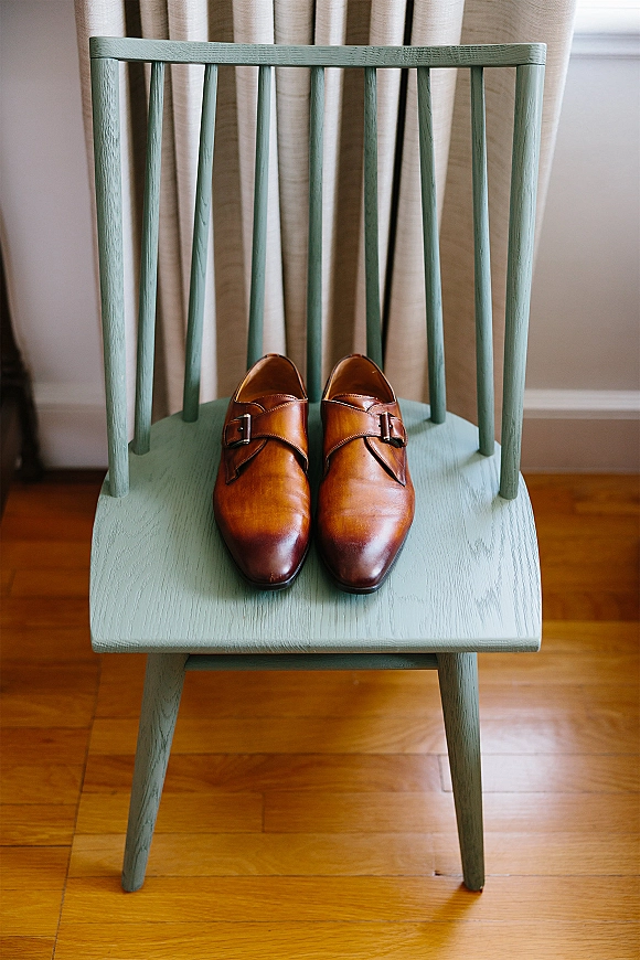 Groom shoes, brown leather dress shoes with double monk strap buckles resting on a wooden chair by a window with curtains