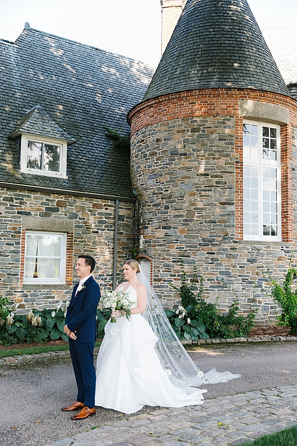 Wedding first look as bride taps groom’s shoulder, bouquet in hand, cathedral veil trailing on cobblestones by an ivy stone manor house