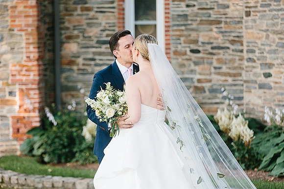 Wedding kiss portrait of bride and groom kissing, her embroidered cathedral veil and white rose bouquet against a stone and brick wall backdrop