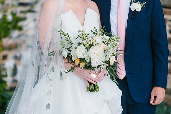 Wedding couple portrait with bride holding a white-and-green bouquet as groom in navy suit with pink tie stands by a stone wall