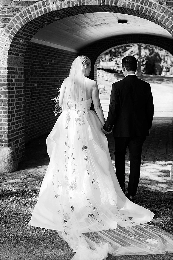 Couple portrait of newlyweds holding hands, bride’s embroidered veil and train flowing as they walk under a brick archway tunnel