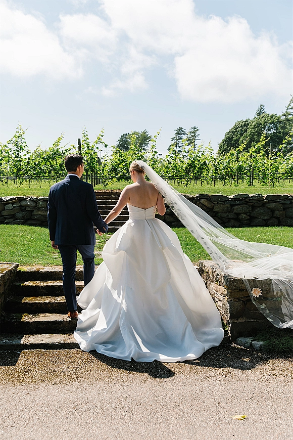 Couple portrait of newlyweds holding hands walking up stone steps in a vineyard, bride’s cathedral veil and dress train flowing behind
