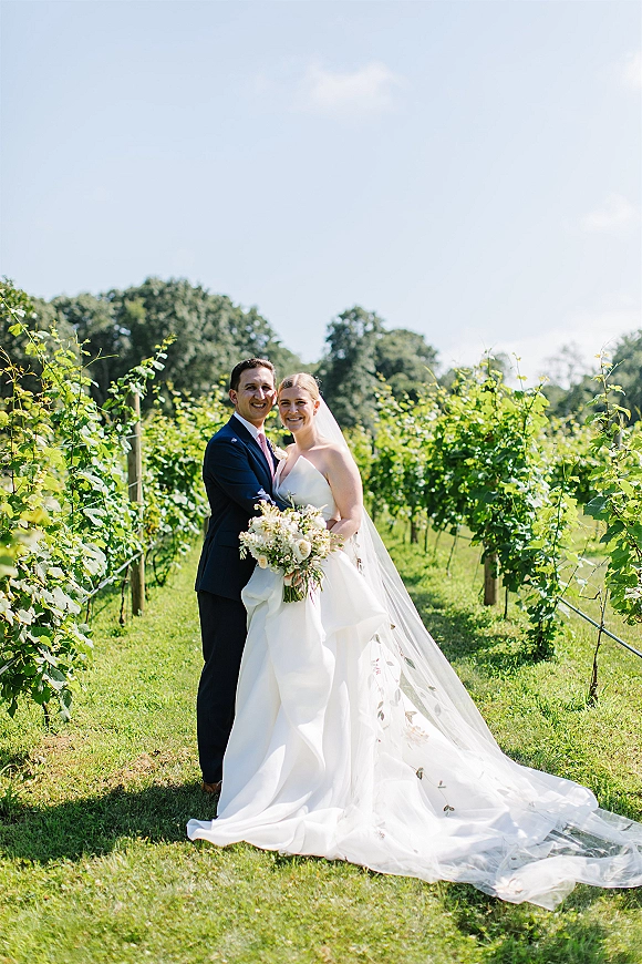 Couple portrait at a vineyard wedding, bride in strapless dress with cathedral veil holding bouquet, groom in navy suit under blue sky
