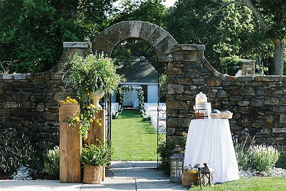 Ceremony entrance decor with a wrought iron gate framed by greenery garland, lemon branches, and urn florals along a garden aisle