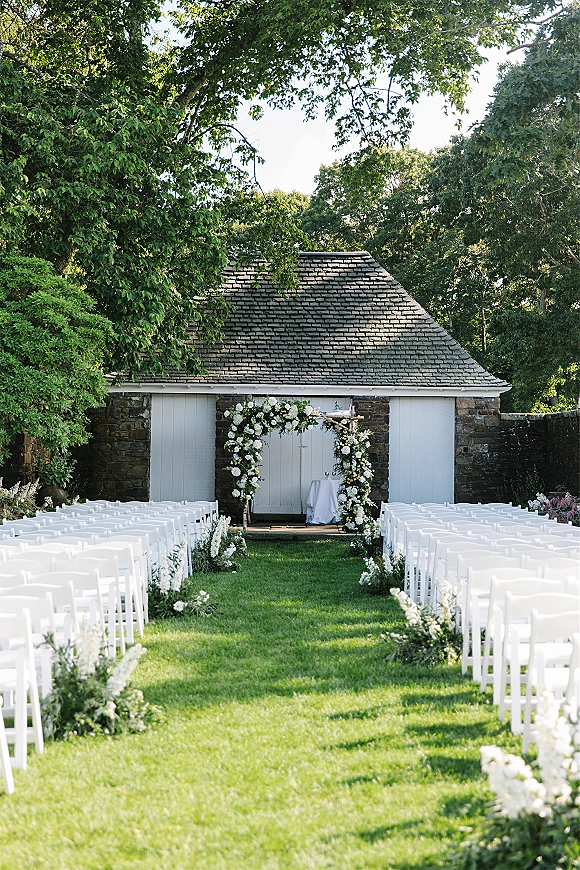 Outdoor ceremony setup with white folding chairs facing a floral arch, aisle lined with white flowers and greenery on a lawn by a stone building