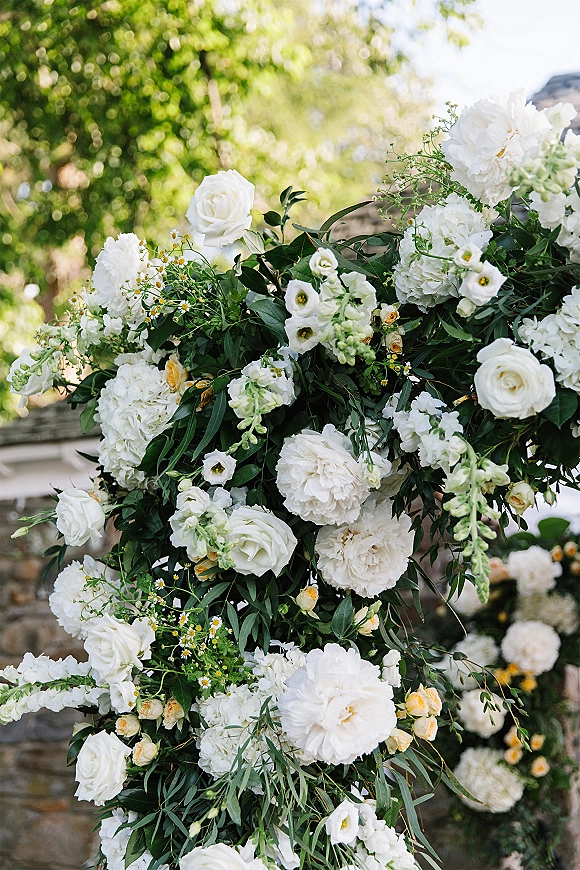 Wedding floral arch of white roses, peonies, and lisianthus with trailing greenery, set against garden trees and a stone wall