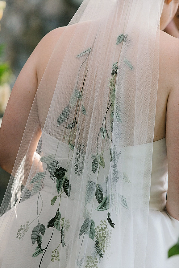 Bridal veil with floral embroidery cascading over a strapless wedding dress, set against softly blurred outdoor greenery in back view
