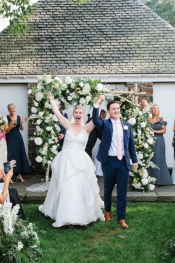 Wedding recessional as newlyweds walk back up aisle with raised hands, bride lifting bouquet overhead under a floral arch on grass lawn