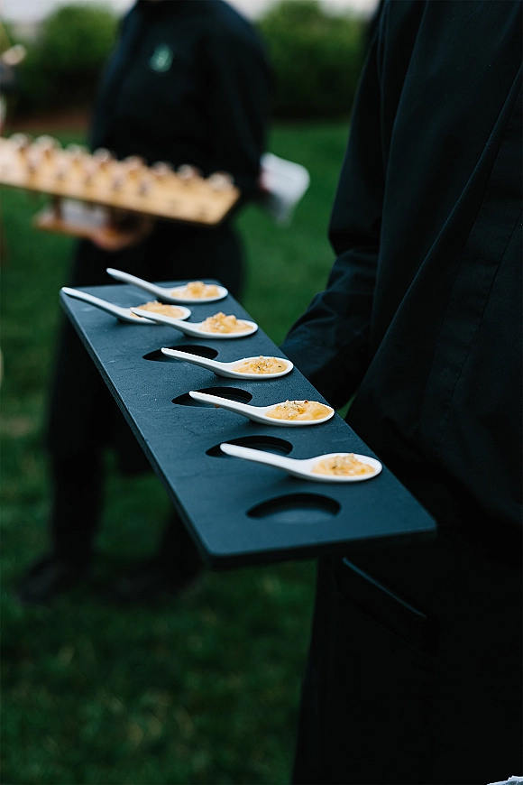 Passed appetizers on a serving tray with ceramic tasting spoons, carried by catering staff in black uniform on a grassy lawn