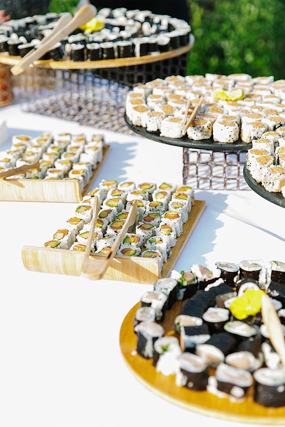 Sushi catering spread with sushi rolls and chopsticks on bamboo boats and cake stands, with lemon wedges on a white linen table outdoors