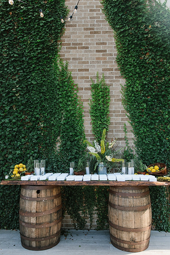 Escort card table with wedding escort cards on a long wooden top, wine barrels, greenery garland, candles, lemons, and ivy brick wall backdrop