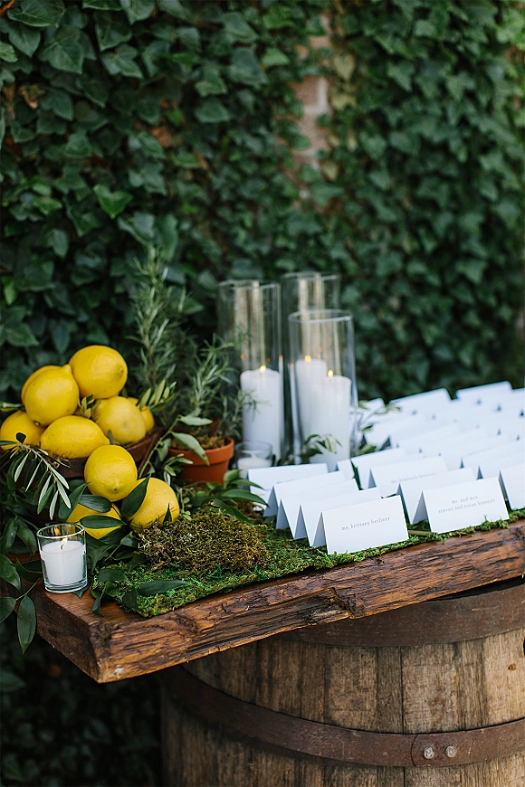 Wedding escort cards arranged on a rustic wooden table with lemons, greenery, rosemary, moss runner and candles against an ivy wall backdrop