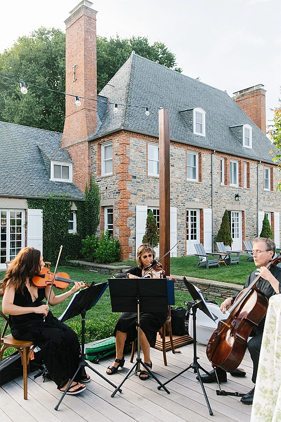 Wedding musicians performing as a string quartet wedding with violin and cello at music stands on a lawn beside a stone house under string lights