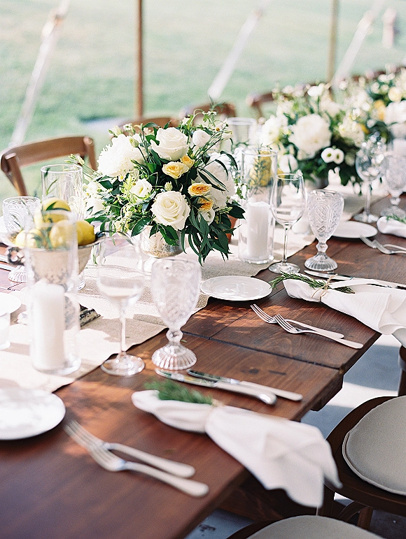 Reception tablescape on a wood farm table wedding setup with white rose centerpieces, taper candles, lemons, and greenery under an outdoor tent