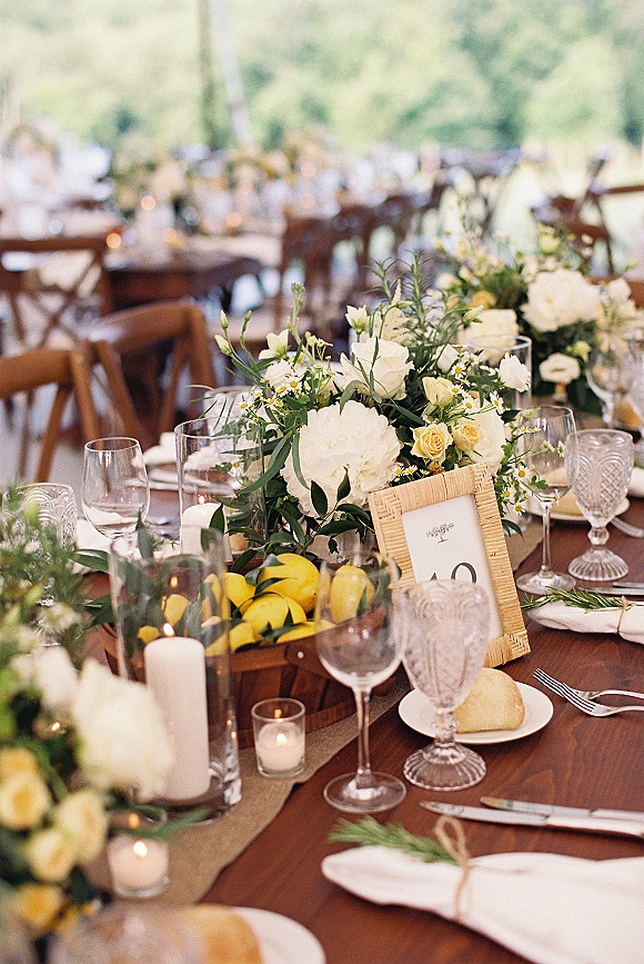 Reception tablescape with white floral centerpieces and greenery garland, lemons and candles on a long banquet table under an outdoor tent