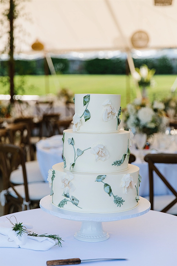 Wedding cake with tiered white frosting and sugar flowers, green foliage details on a stand at a white tent reception with tables and chairs