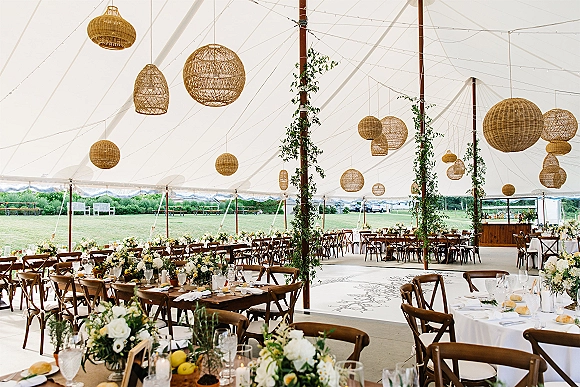 Tent wedding reception under a white sailcloth canopy with rattan pendant lanterns, string lights, and long farmhouse tables on a lawn