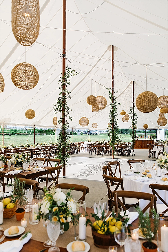 Tent reception decor under a white sailcloth tent with wicker pendant lights, string lights, greenery garlands, and candlelit tables on grass lawn