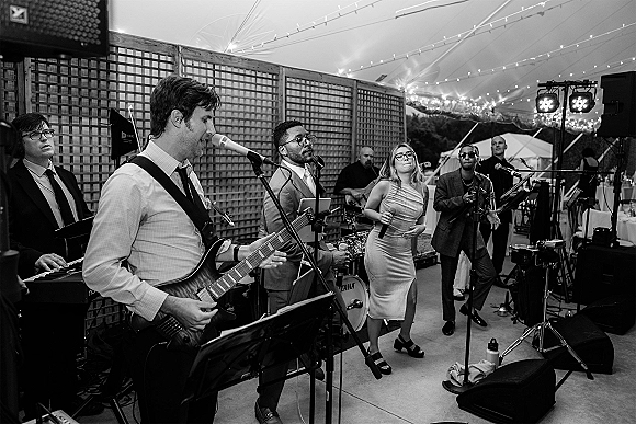 Wedding band playing live wedding music with microphones and electric guitar under string lights on an outdoor patio beneath a tent canopy