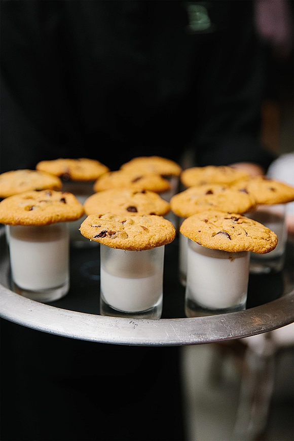 Wedding dessert shots of cookie and milk in shot glasses with chocolate chip cookies on a serving tray, held by a server at reception
