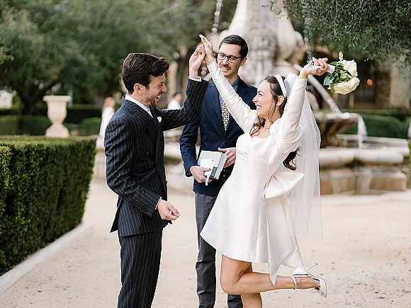 Ceremony moment during wedding ring exchange as bride lifts bouquet beside groom in pinstripe suit, with a stone fountain behind them