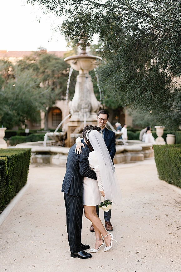 Wedding ceremony moment as bride hugs groom, holding a white bouquet in a courtyard garden by a stone fountain with arches behind them