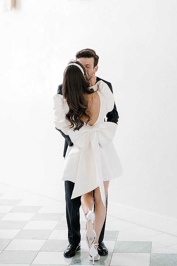 Wedding kiss portrait of bride and groom kissing as he hugs her; she wears a short white open-back dress with bow against a white wall
