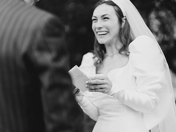 Wedding vows as the bride reading vows from a vow book, smiling in a long sleeve dress and veil with trees in the background